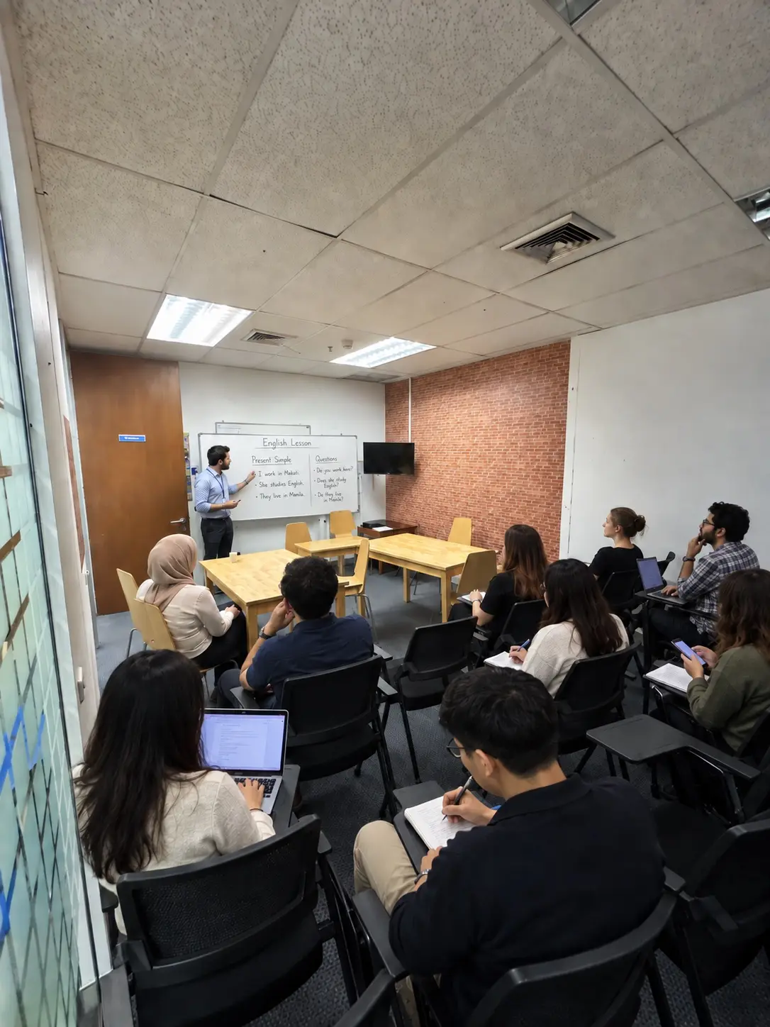 An English class at the school: the teacher explains the present simple on the whiteboard while students follow with notebooks and laptops