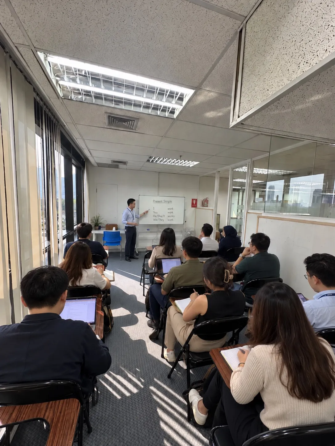A class at the school in a room by the windows: the teacher writes on the whiteboard while adult students take notes